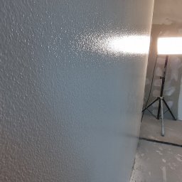 BauTech - Świeżo pomalowana szara ściana z widoczną fakturą, oświetlona lampą budowlaną w trakcie prac wykończeniowych.