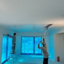 BauTech - Dwóch malarzy podczas pracy w jasnym, przestronnym wnętrzu. Okna zabezpieczone folią, widoczne narzędzia malarskie i drabina.