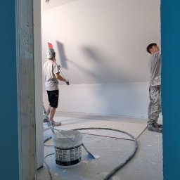 BauTech - Dwóch malarzy w trakcie malowania białej ściany w pomieszczeniu o skośnym suficie, widok przez framugę drzwi, na podłodze wiadro z farbą i wąż od agregatu.