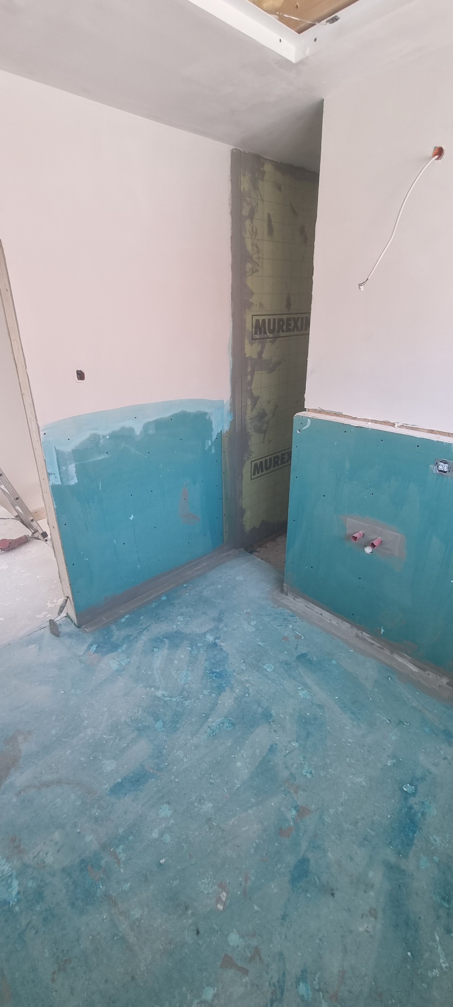 Przygotowanie ścian i podłogi w łazience przed położeniem płytek, widoczne płyty kartonowo-gipsowe z nałożoną warstwą hydroizolacji w kolorze turkusowym oraz zabezpieczona folią hydroizolacyjną...