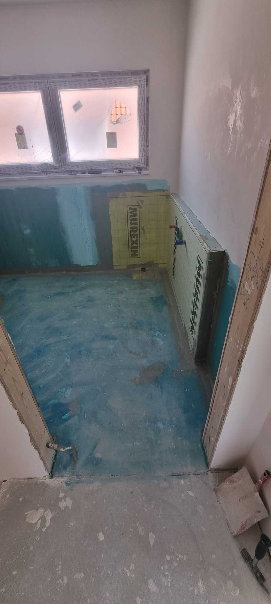 Przygotowanie łazienki do położenia płytek: hydroizolacja w kolorze niebieskim na podłodze i ścianach, żółta mata uszczelniająca Murexin na ścianach, widoczne rury instalacji wodnej, surowy stan...