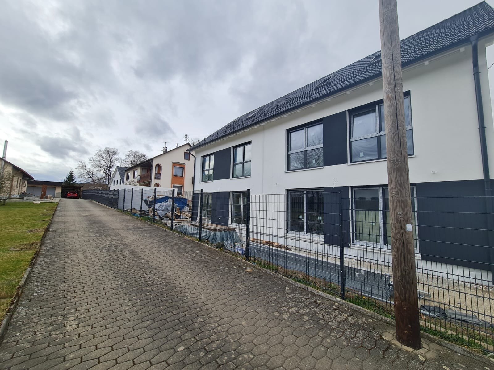 Nowoczesny dom w Szczecinie z białą elewacją i grafitowymi akcentami, widoczny z boku posesji, z elementami ogrodzenia panelowego i brukowaną drogą dojazdową.