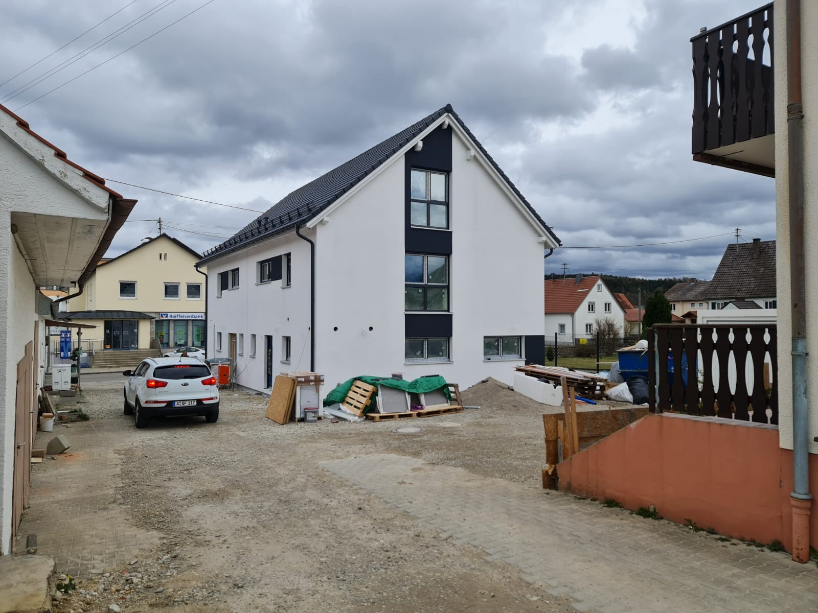 Dwukondygnacyjny dom z białymi ścianami i czarnymi akcentami, z ciemną dachówką, widoczny z boku, na placu budowy z materiałami budowlanymi i zaparkowanym samochodem.