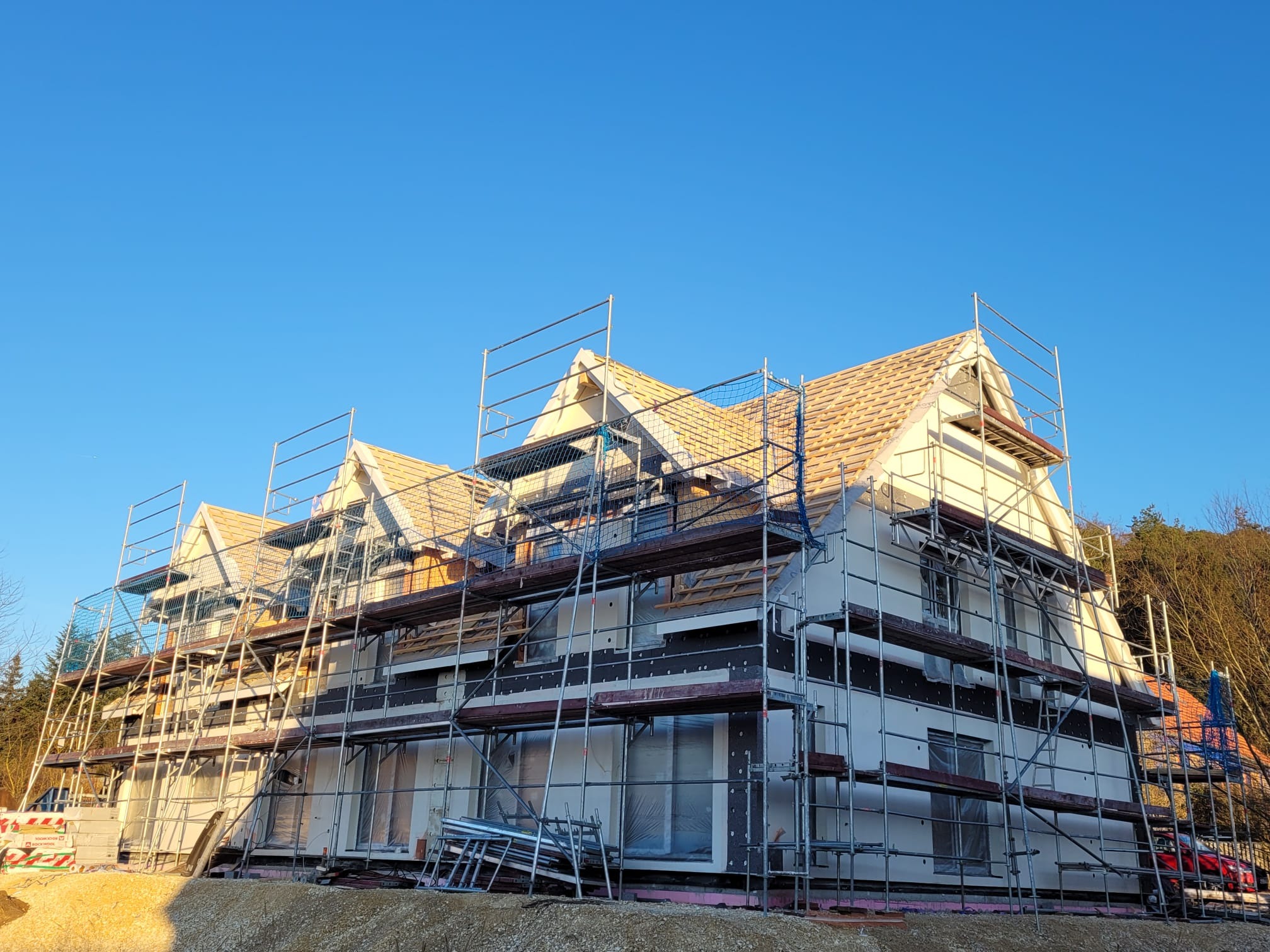 Wielopiętrowy dom w trakcie budowy, otoczony rusztowaniami, z widoczną konstrukcją dachu i częściowo otynkowaną elewacją, na tle błękitnego nieba.