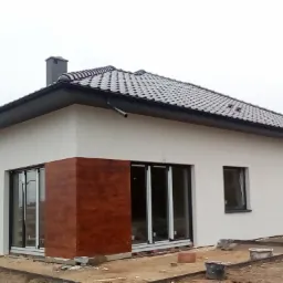 Nowoczesny dom jednorodzinny z jasną elewacją i elementami dekoracyjnymi imitującymi drewno, widoczne okna z czarnymi ramami i przygotowany teren wokół budynku.