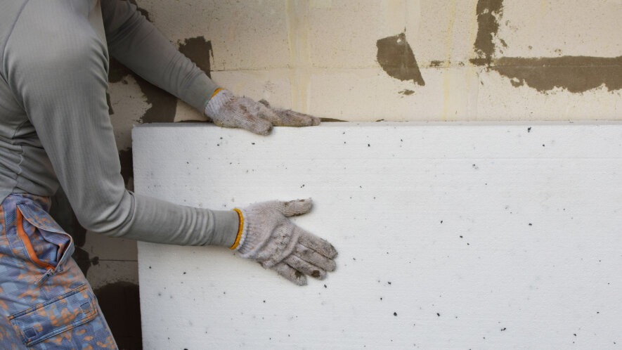 Pracownik w ubrudzonych rękawicach dociska białą płytę styropianową do surowej ściany z pustaków, widoczne ślady zaprawy.