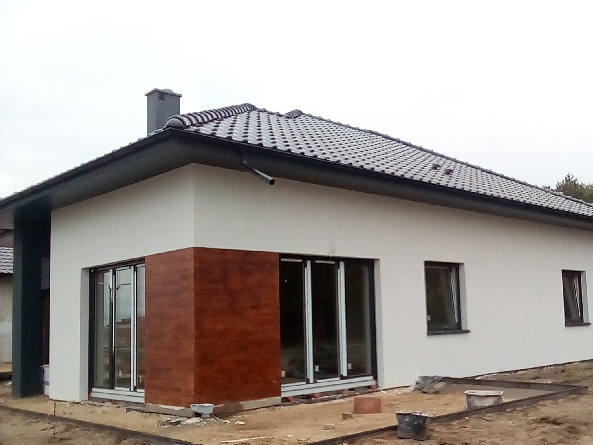 Nowoczesny dom jednorodzinny z jasną elewacją i elementami dekoracyjnymi imitującymi drewno, widoczne okna z czarnymi ramami i przygotowany teren wokół budynku.