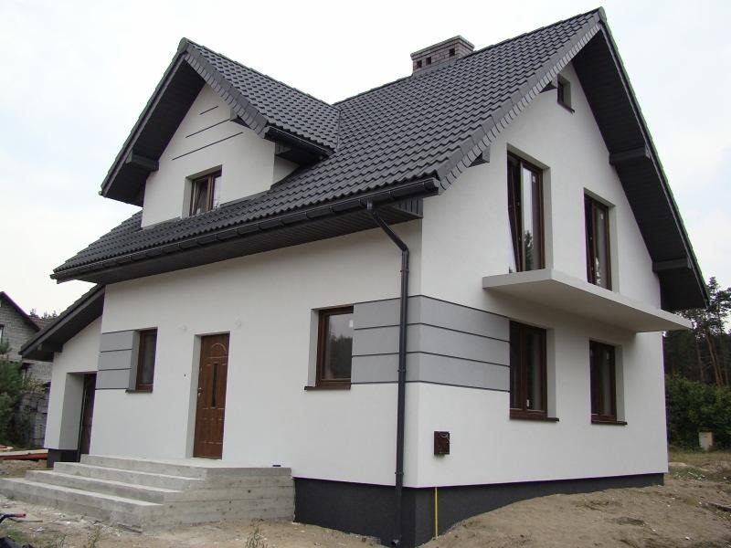 Dwukondygnacyjny dom z białą elewacją, ciemną dachówką i szarymi akcentami, widoczne betonowe schody wejściowe i fragment niezagospodarowanego terenu.