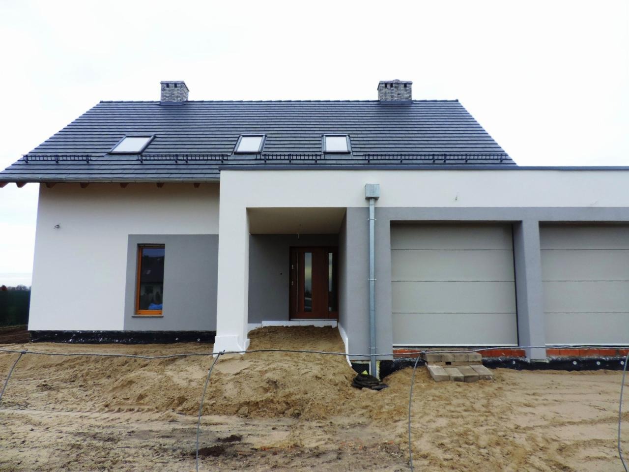 Nowoczesny dom jednorodzinny z szarą dachówką i jasną elewacją w trakcie prac wykończeniowych, widoczne niedokończone otoczenie i piasek.