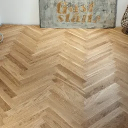 Ułożony parkiet w jodełkę w jasnym odcieniu drewna, widoczne białe ściany w trakcie wykończenia, dekoracyjna lampa z wikliny oraz obraz z napisem w tle.