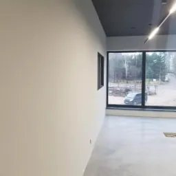 Świeżo pomalowane wnętrze z dużym oknem z widokiem na zewnątrz, szare ściany i sufit, surowy beton na podłodze, widoczny fragment instalacji w podłodze.