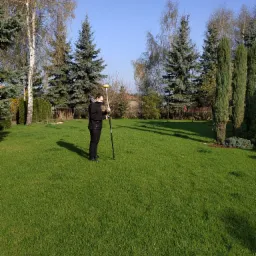 Geodeta w czarnej kurtce mierzy teren na trawniku z drzewami iglastymi w tle, używając tyczki geodezyjnej z żółtą końcówką.