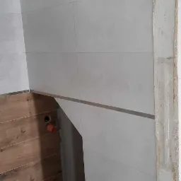 Ściana łazienki w trakcie wykańczania: szare płytki ceramiczne z metaliczną listwą, drewnopodobne panele na ścianie obok, widoczne instalacje hydrauliczne i surowy fragment ściany z resztkami zaprawy.