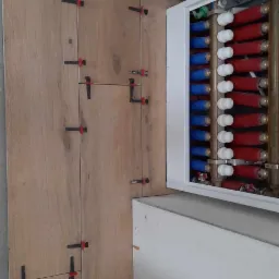 Montaż płytek imitujących drewno na ścianie, z widocznymi czerwono-czarnymi krzyżykami dystansowymi, obok skrzynki z rurami w kolorach czerwonym i niebieskim.