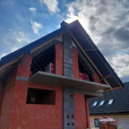 Budowa domu jednorodzinnego w trakcie wznoszenia ścian i konstrukcji dachu, widoczna więźba dachowa, bloczki ceramiczne i betonowe, dachówka na sąsiednim budynku w tle.