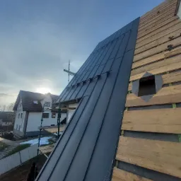 Częściowo pokryty dach blachą na rąbek stojący, widoczna konstrukcja drewniana i otwór wentylacyjny, w tle budynek mieszkalny i rusztowanie.