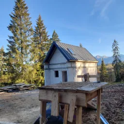 Surowy budynek w trakcie budowy z ciemnoszarym dachem z blachy na tle gór i lasu, widok zza drewnianego stołu roboczego.