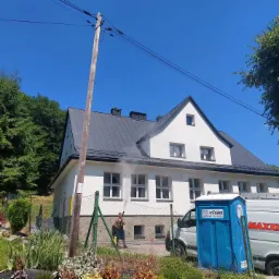 Czyszczenie elewacji domu z szarym dachem za pomocą myjki ciśnieniowej, widoczny pracownik w krótkich spodenkach i przenośna toaleta w tle.