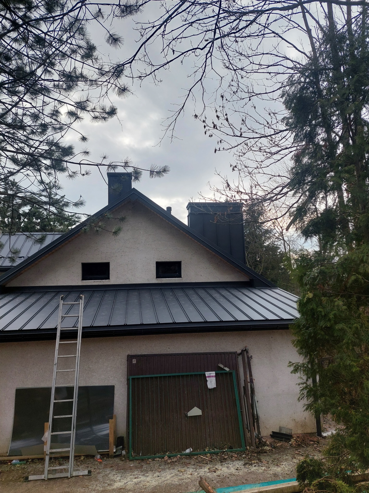 Dom z nowym, ciemnym, metalowym pokryciem dachowym, widoczne kominy i drabina oparta o ścianę budynku. W tle drzewa i pochmurne niebo.