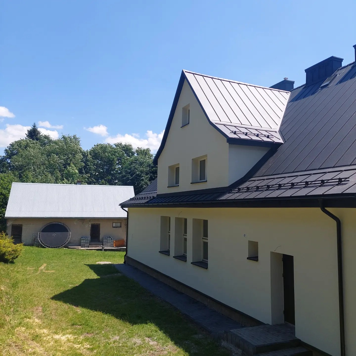 Nowoczesny dom z ciemnoszarym dachem z blachy na tle zielonego ogrodu i błękitnego nieba.