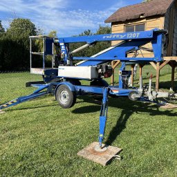 PHU JAN SAWICKI - Niebieski podnośnik koszowy Nifty 120T na trawie, z wysuniętymi podporami na drewnianych podkładkach. W tle domek na drzewie i zieleń.