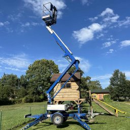 PHU JAN SAWICKI - Niebieski podnośnik koszowy na trawniku, z wysięgnikiem skierowanym ku górze, na tle błękitnego nieba i drewnianego domku dla dzieci. Widoczne podpory stabilizujące.
