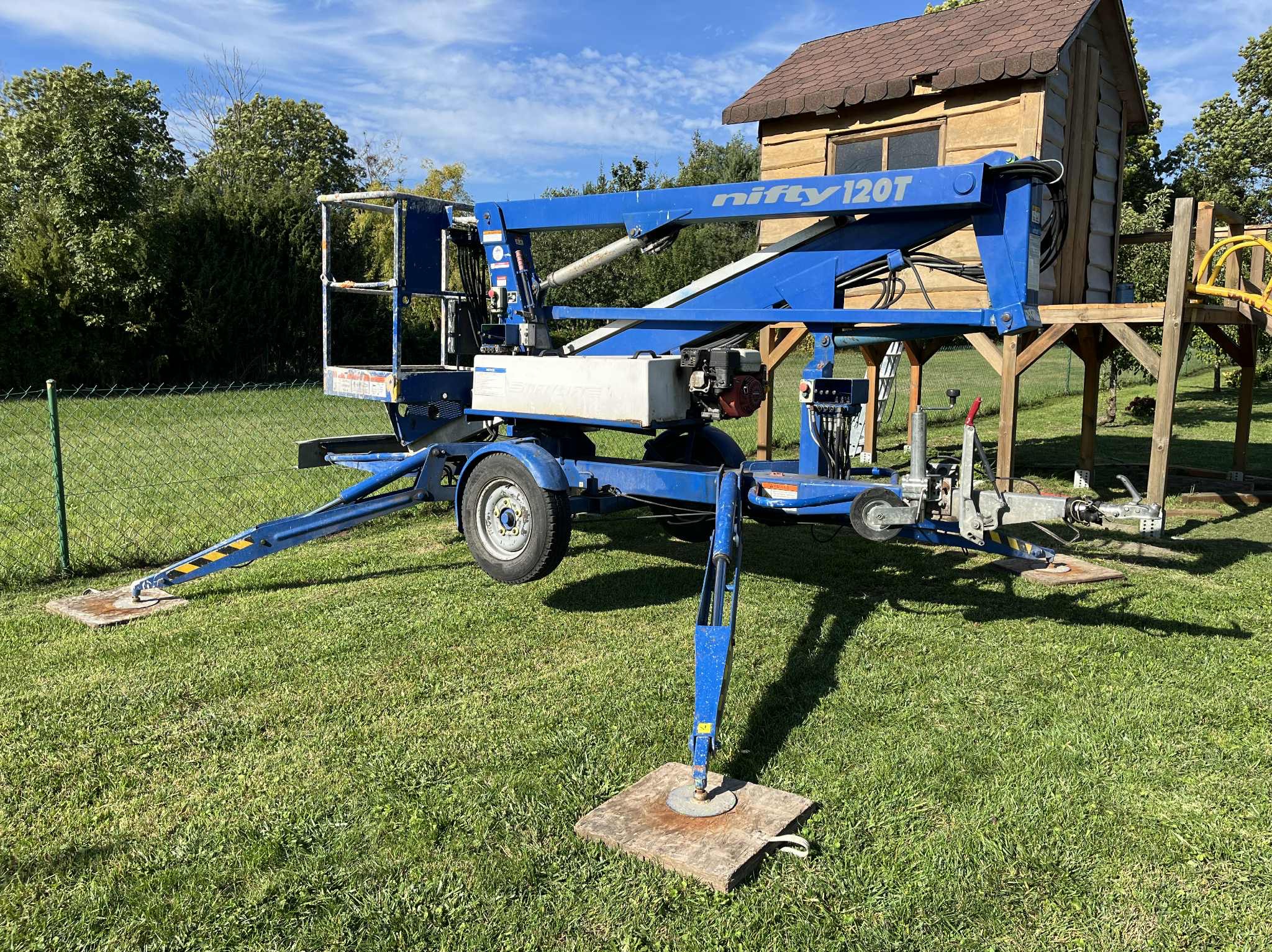 Niebieski podnośnik koszowy Nifty 120T na trawie, z wysuniętymi podporami na drewnianych podkładkach. W tle domek na drzewie i zieleń.