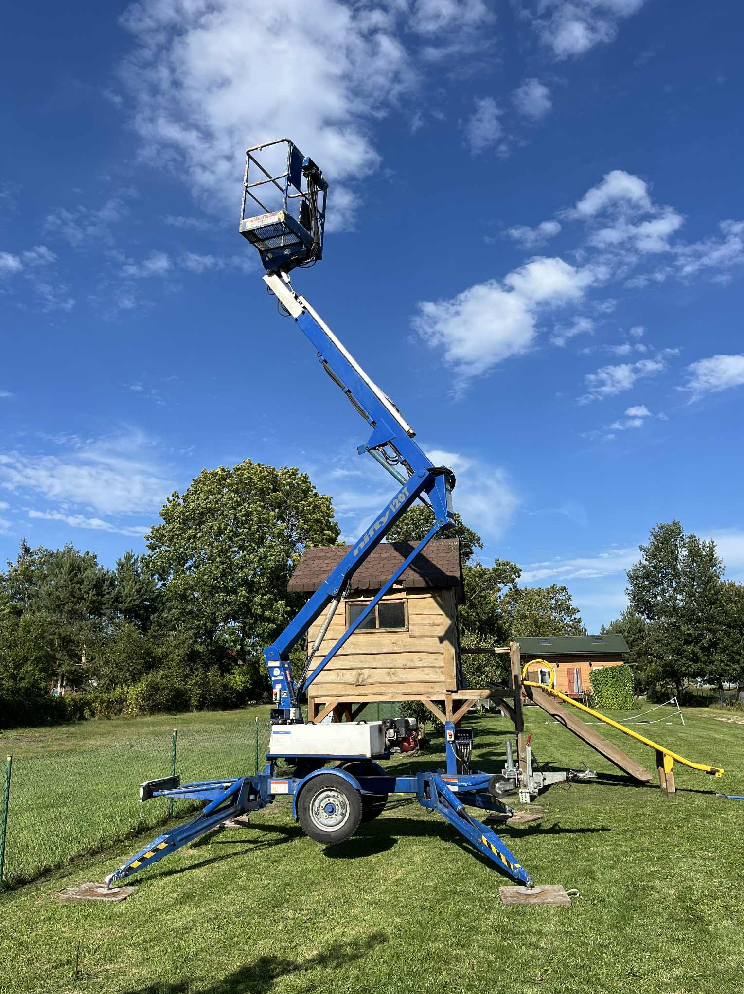 Niebieski podnośnik koszowy na trawniku, z wysięgnikiem skierowanym ku górze, na tle błękitnego nieba i drewnianego domku dla dzieci. Widoczne podpory stabilizujące.