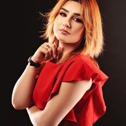 Barbara Bulanda Fotogtrafia - Portret kobiety z rudymi włosami w czerwonej sukience na ciemnym tle, z ręką przy twarzy i pierścionkami na palcach.