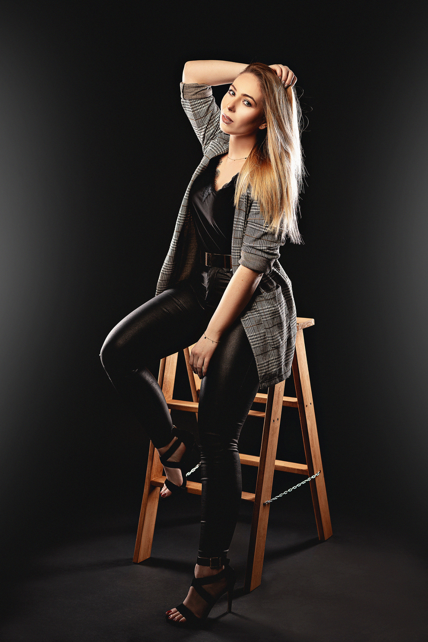 Kobieta w skórzanych spodniach i marynarce w kratę pozuje siedząc na drewnianym stołku na czarnym tle, z ręką uniesioną do włosów.