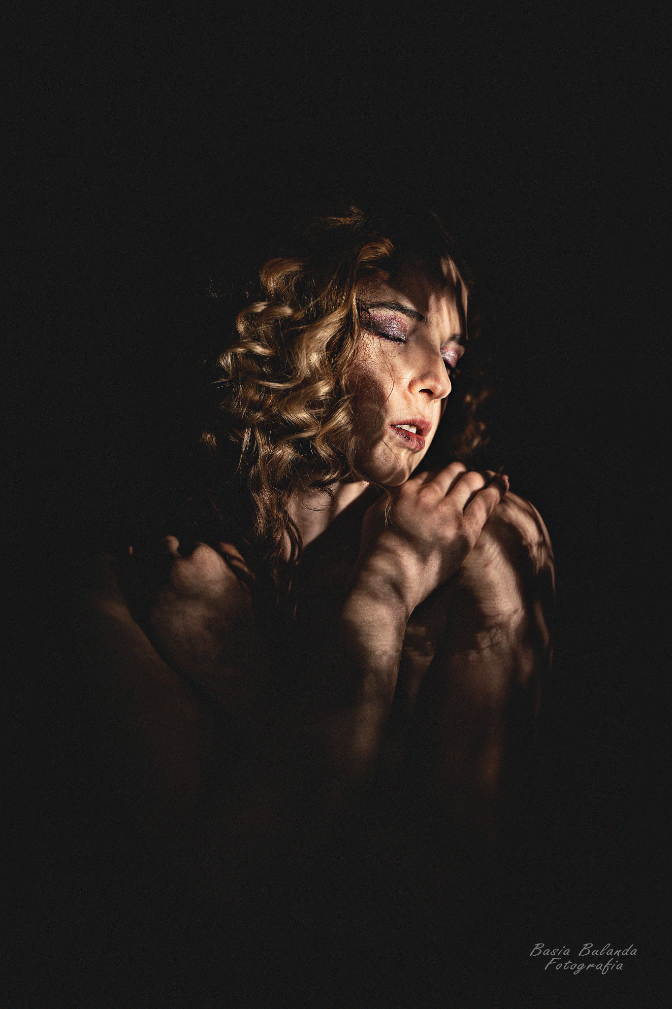 Portret kobiety z zamkniętymi oczami, oświetlonej światłem tworzącym cienie na twarzy i ramionach, z kręconymi blond włosami i brokatowym makijażem oczu.