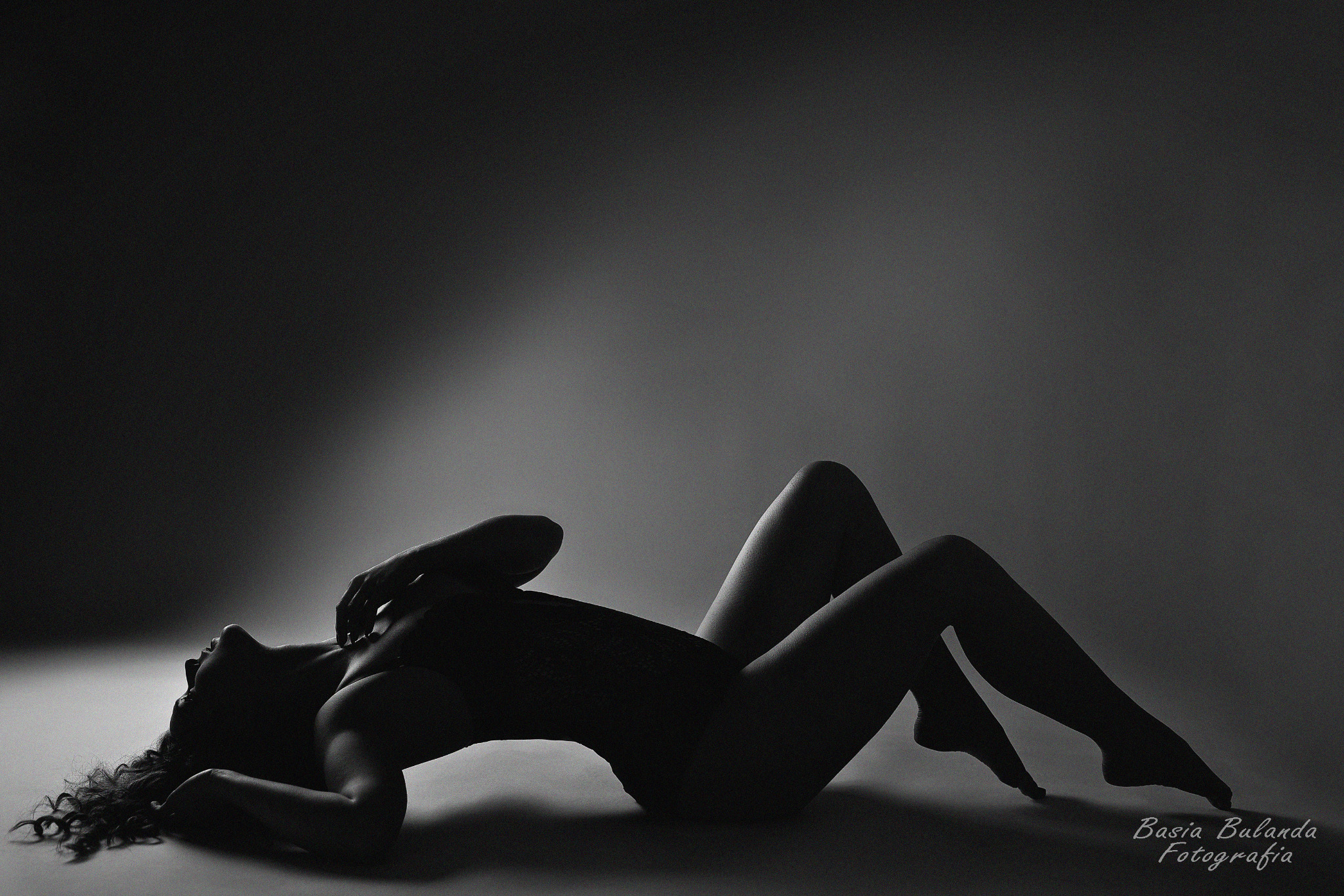 Sylwetka kobiety w bieliźnie, leżącej na podłodze w kontraście światła i cienia, z podpisem 'Basia Bulanda Fotografia' w prawym dolnym rogu.