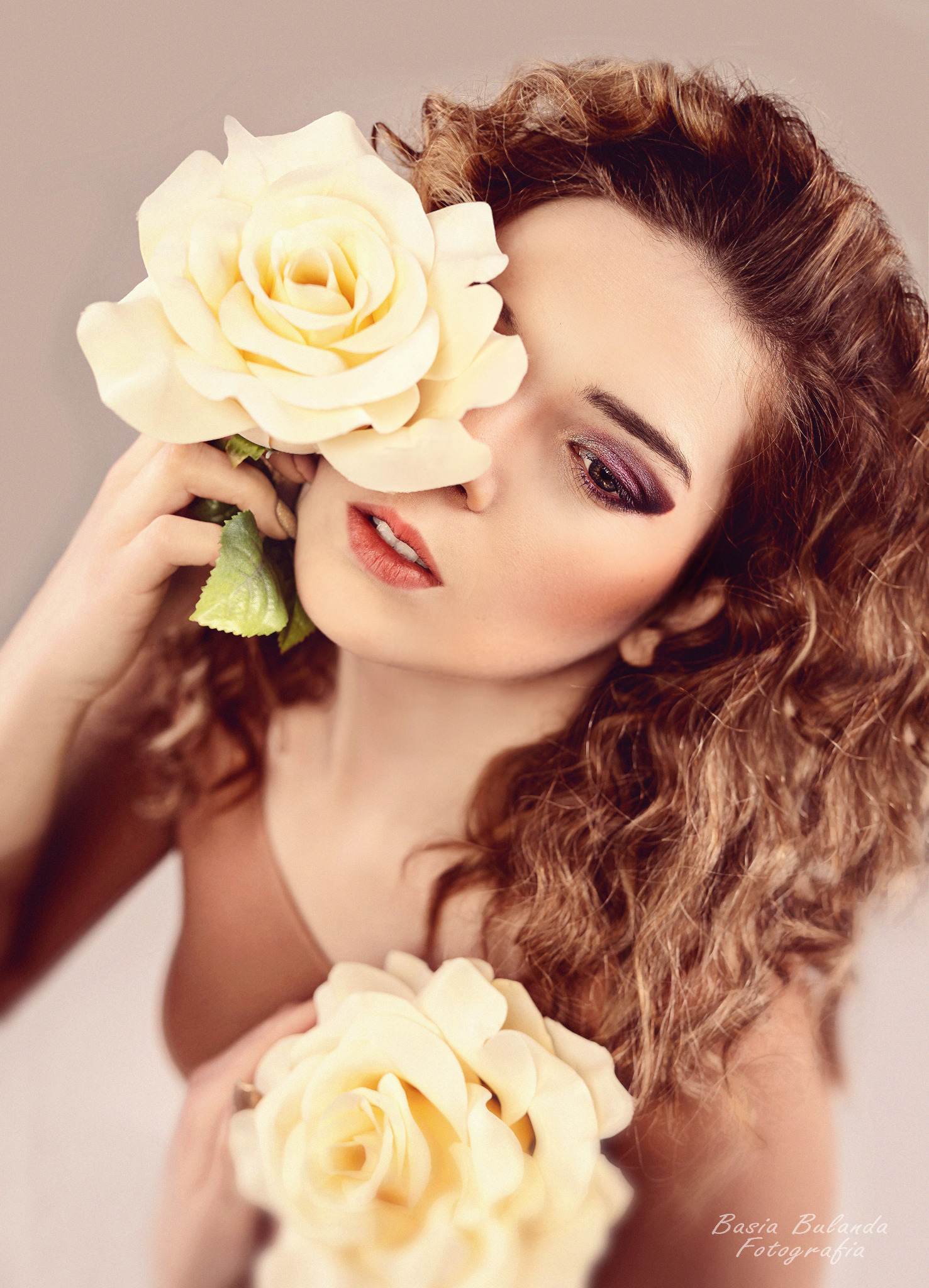 Portret kobiety z burzą loków, zakrywającej oko kremową różą, w subtelnym makijażu.