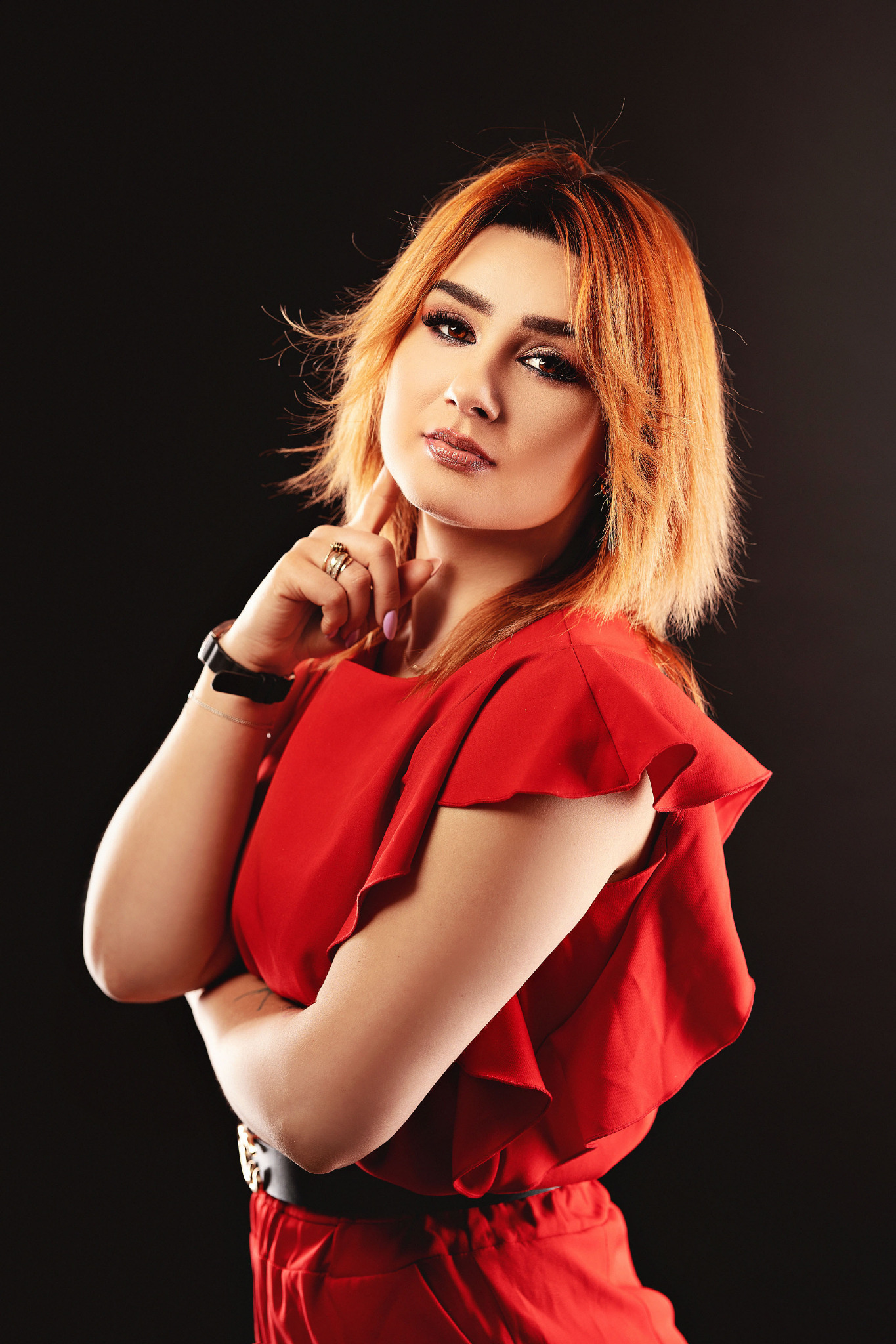 Portret kobiety z rudymi włosami w czerwonej sukience na ciemnym tle, z ręką przy twarzy i pierścionkami na palcach.