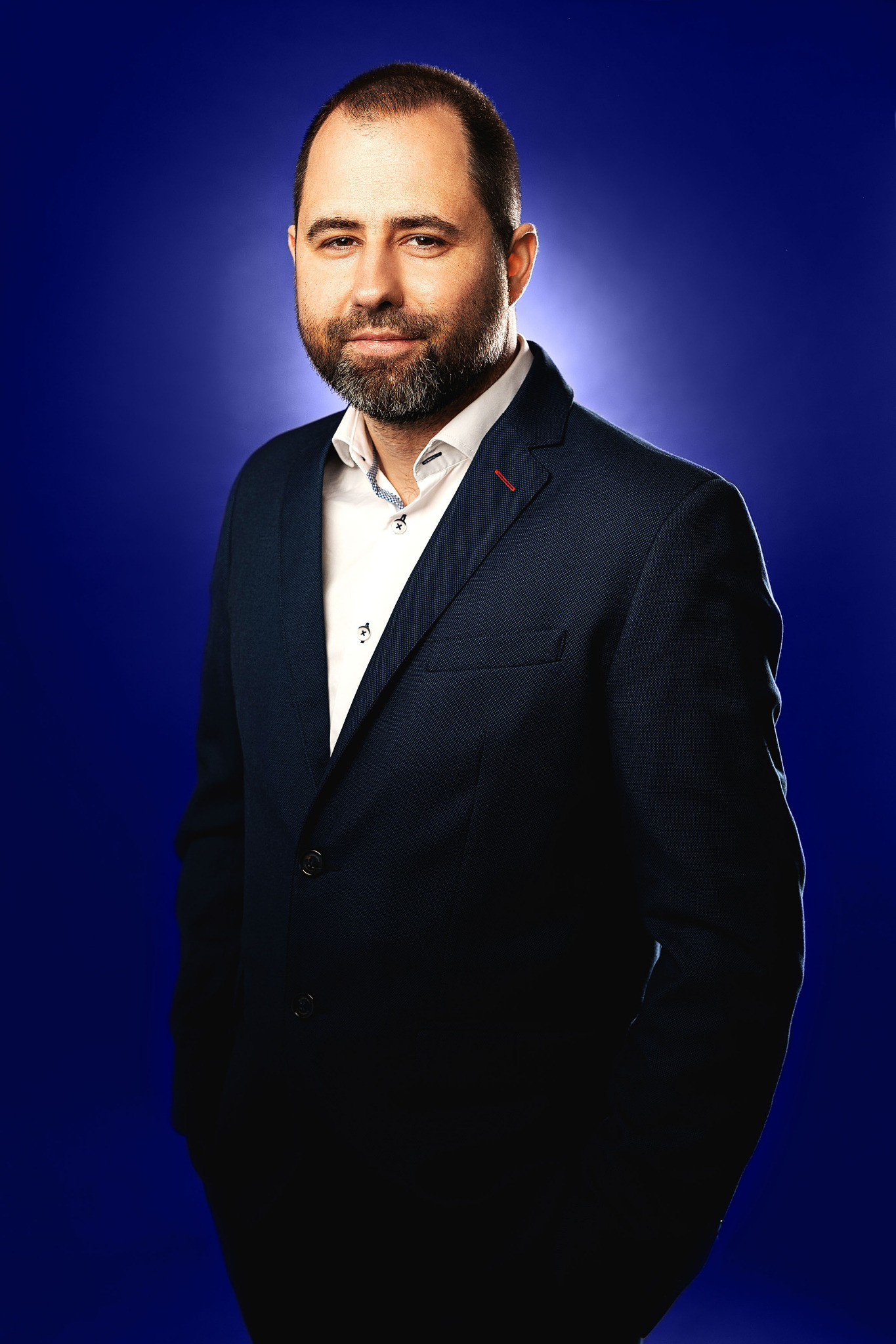 Portret mężczyzny z brodą w ciemnogranatowej marynarce i białej koszuli na jednolitym, ciemnoniebieskim tle.