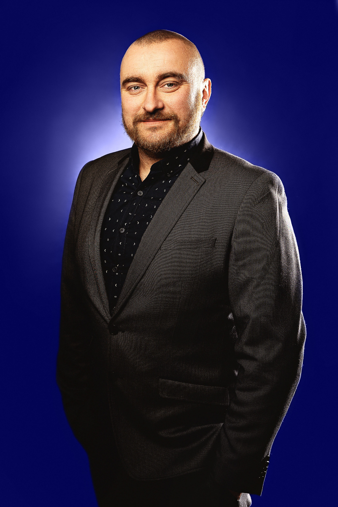 Portret mężczyzny w szarym garniturze i wzorzystej koszuli na jednolitym, ciemnoniebieskim tle, z delikatnym uśmiechem i brodą.