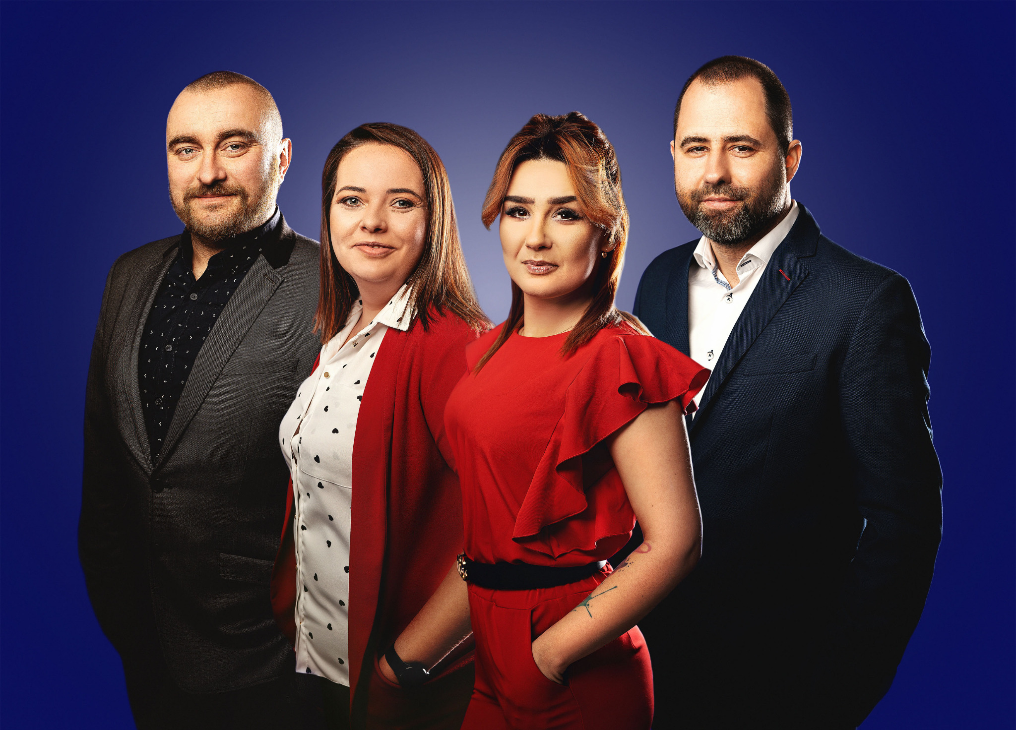 Portret grupowy: dwóch mężczyzn w garniturach i dwie kobiety w czerwonych stylizacjach na ciemnoniebieskim tle.