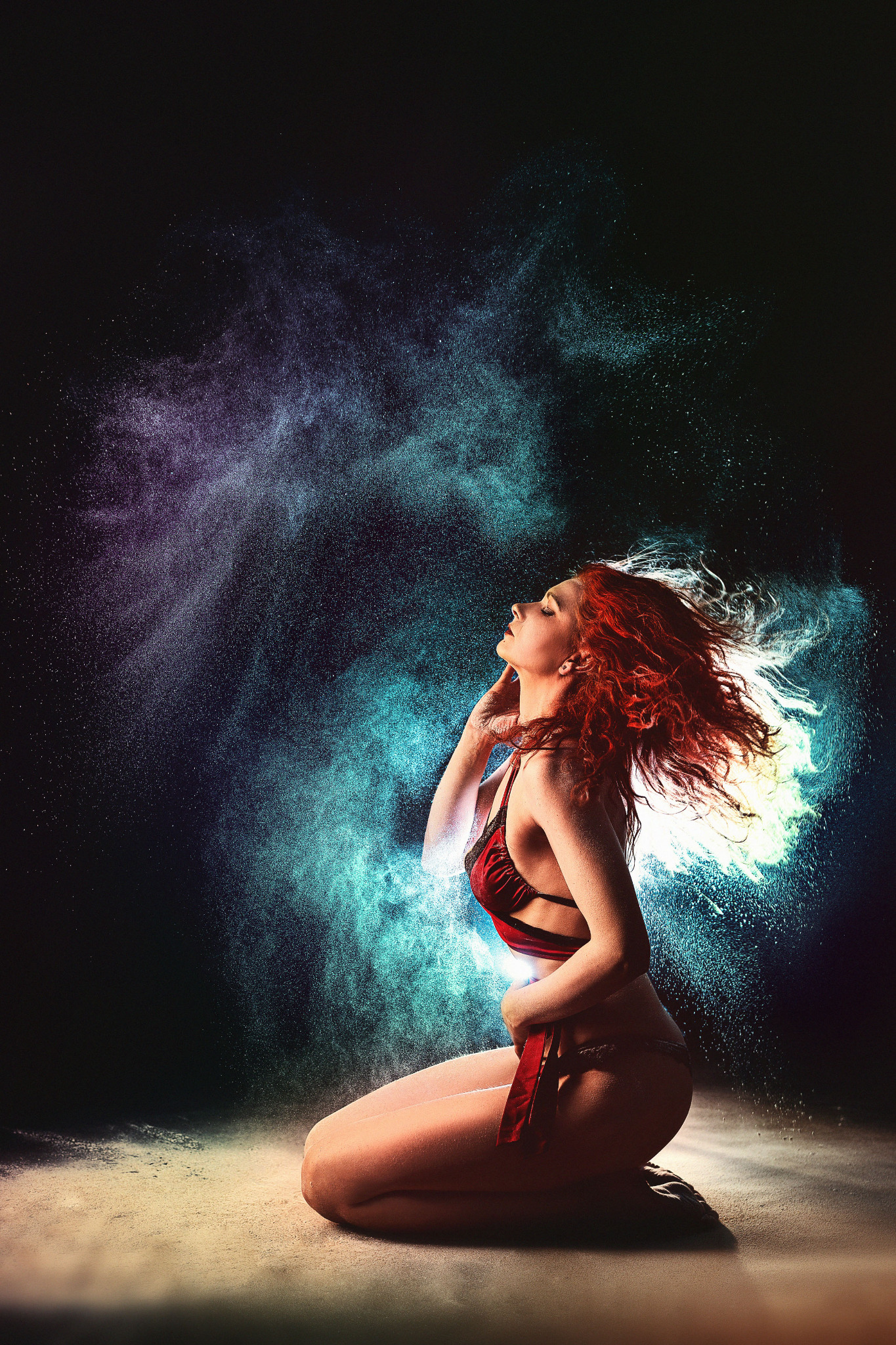 Kobieta z rudymi włosami klęczy w otoczeniu kolorowego pyłu, odchylona głowa do tyłu, dłoń przy szyi, czerwona bielizna.