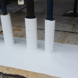 Uszczelnienie przejść rur przez dach płaskim białym materiałem hydroizolacyjnym, widoczne różne średnice rur i nierówności podłoża.