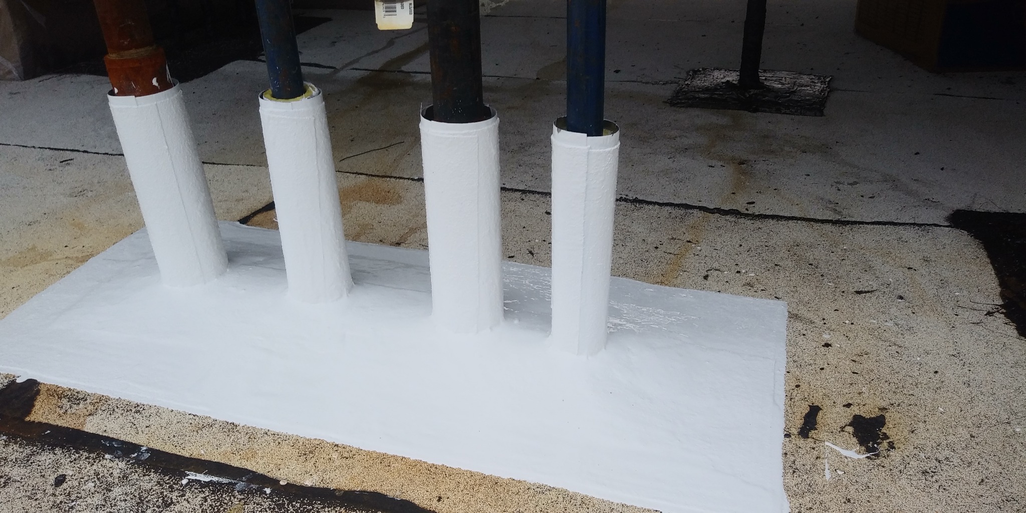 Uszczelnienie przejść rur przez dach płaskim białym materiałem hydroizolacyjnym, widoczne różne średnice rur i nierówności podłoża.