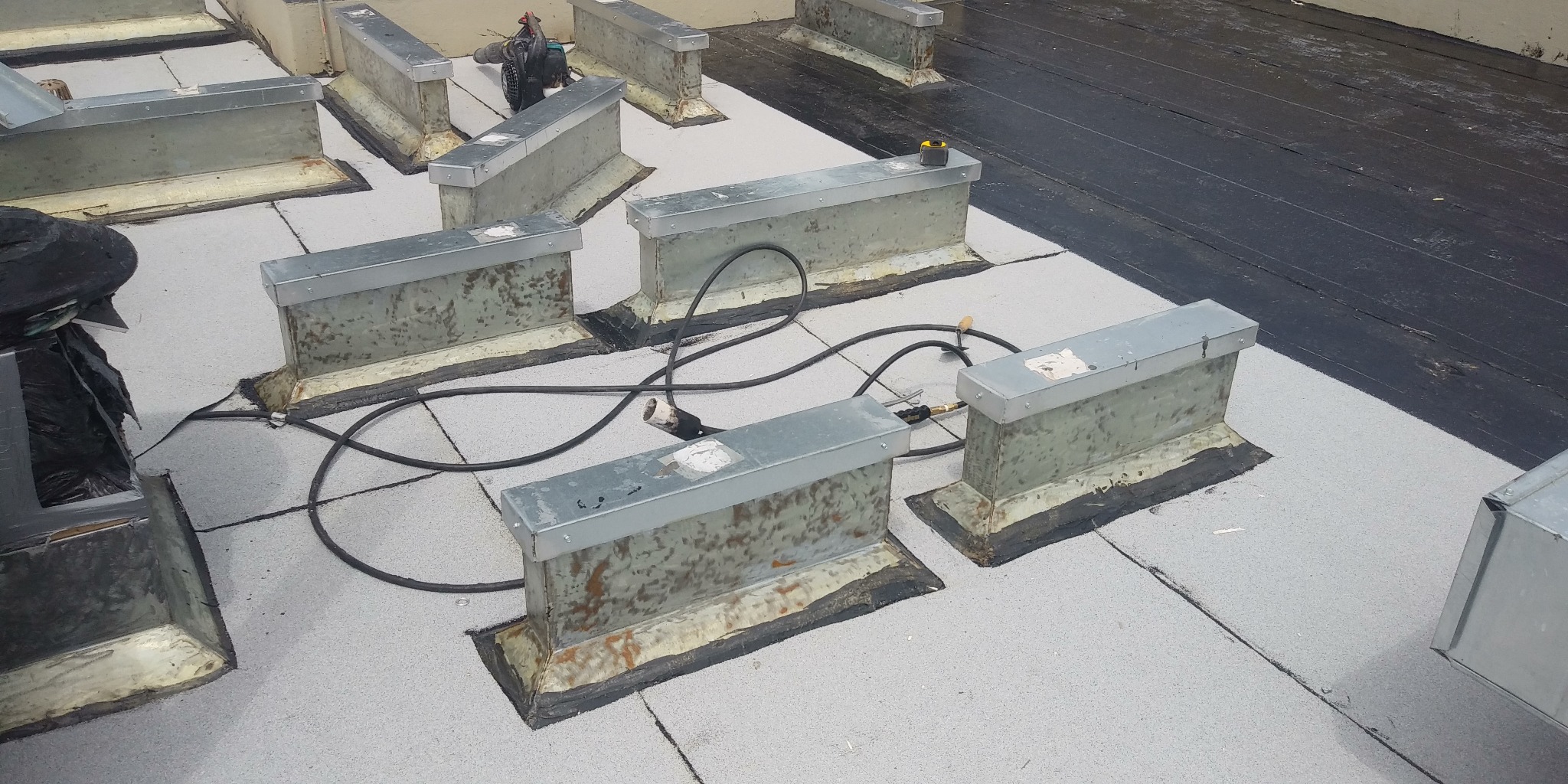 Płaski dach pokryty jasnoszarą papą z widocznymi metalowymi obróbkami kominowymi, czarne kable elektryczne i narzędzia leżące na dachu.