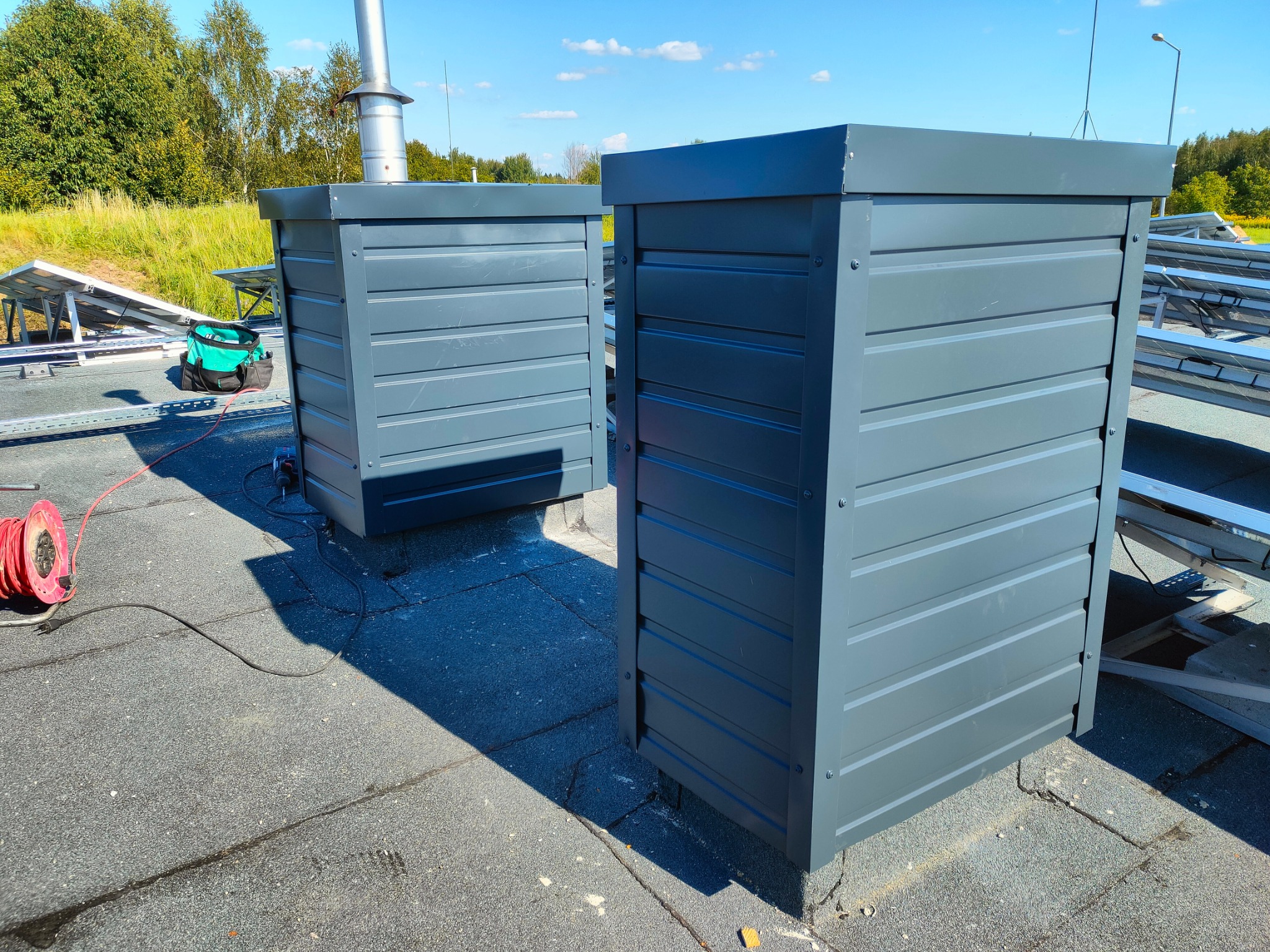 Szare obudowy wentylacyjne na płaskim dachu z papy, widoczne panele słoneczne i narzędzia, instalacja wentylacji na dachu.