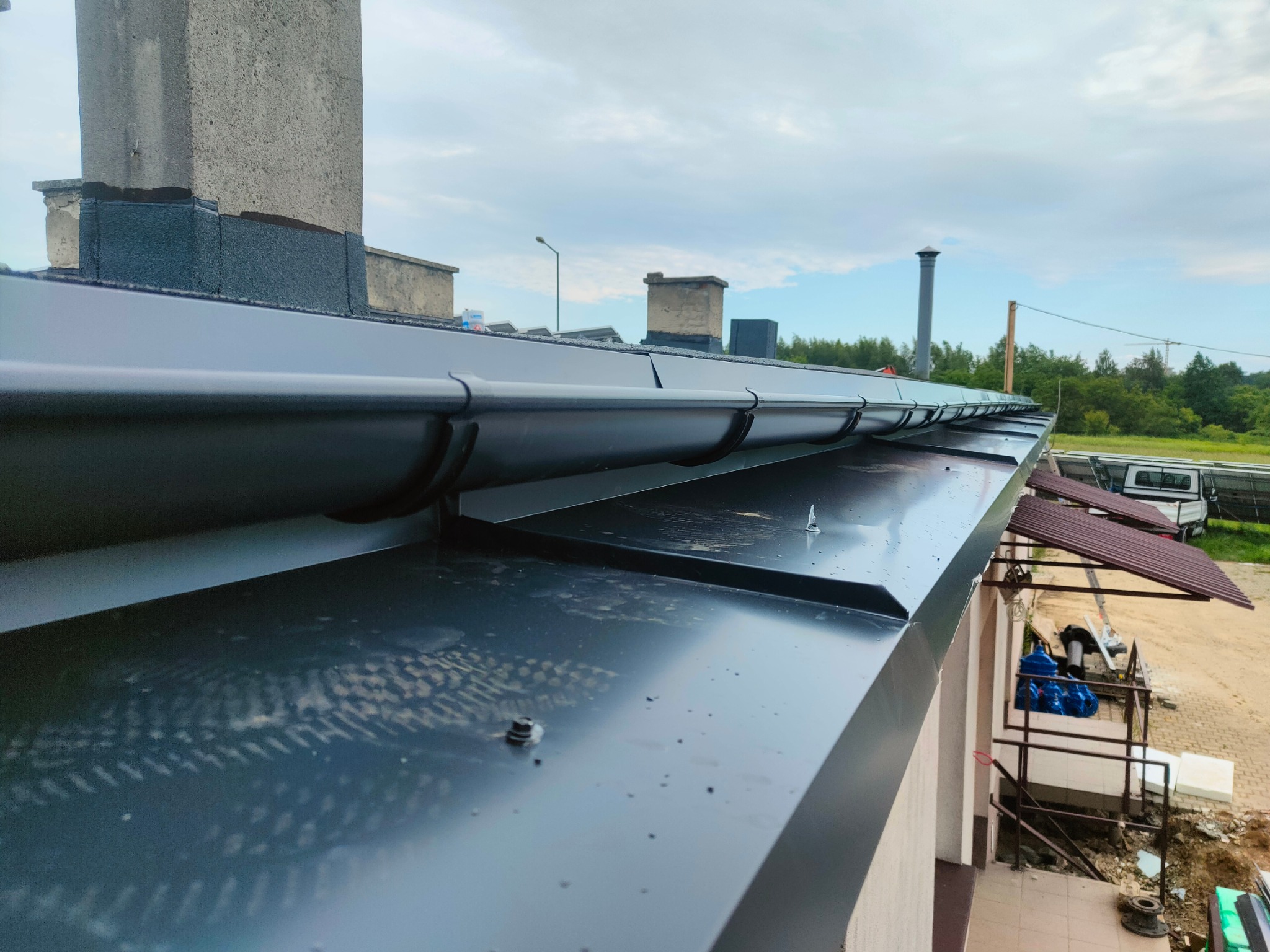 Nowy, błyszczący, czarny dach z orynnowaniem i obróbkami blacharskimi, widok z góry, z kominem w tle i fragmentem terenu posesji po prawej stronie.