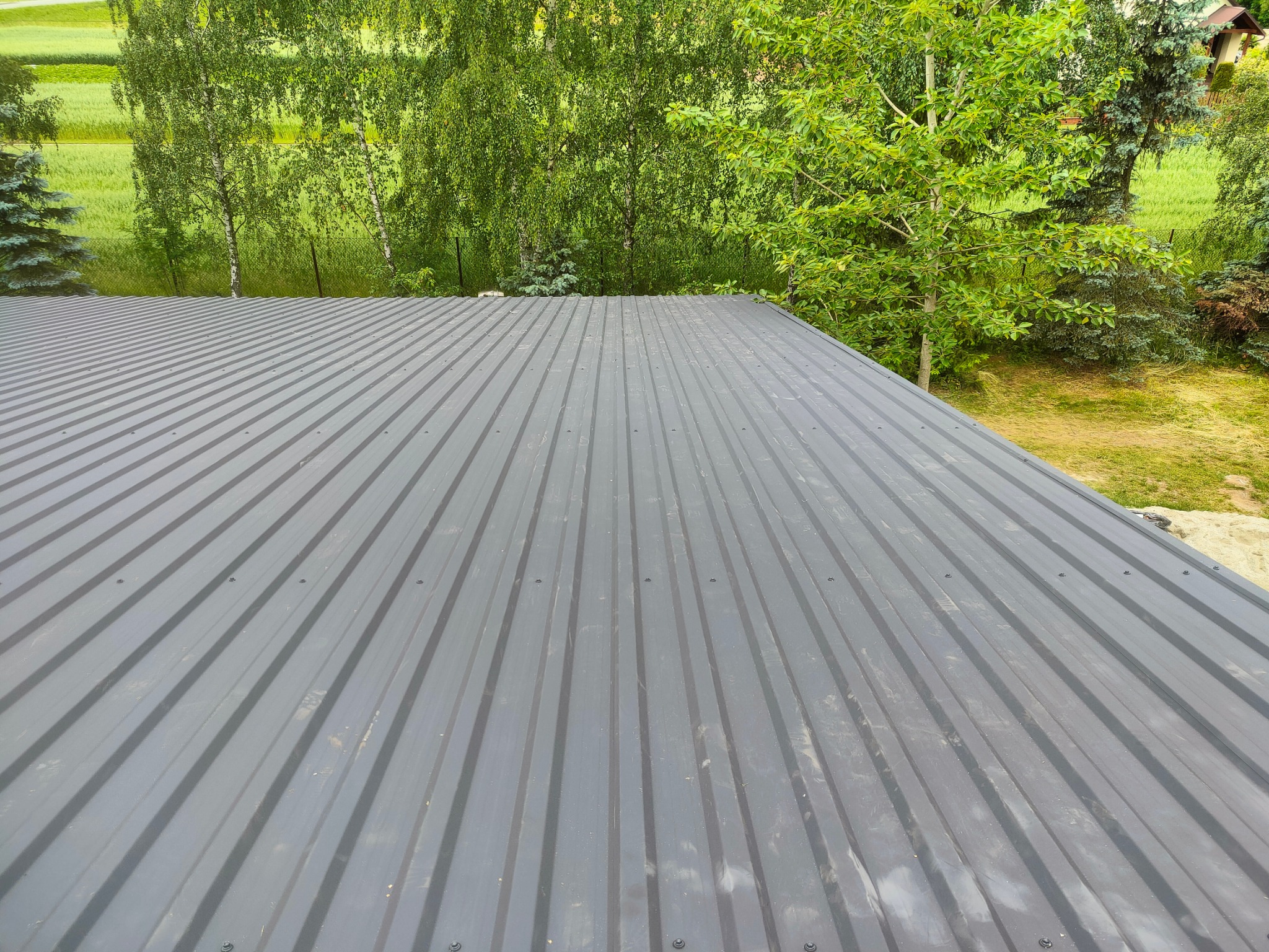 Szary dach z blachy trapezowej, widok z góry na tle zieleni drzew i pola. Nowoczesne pokrycie dachowe.