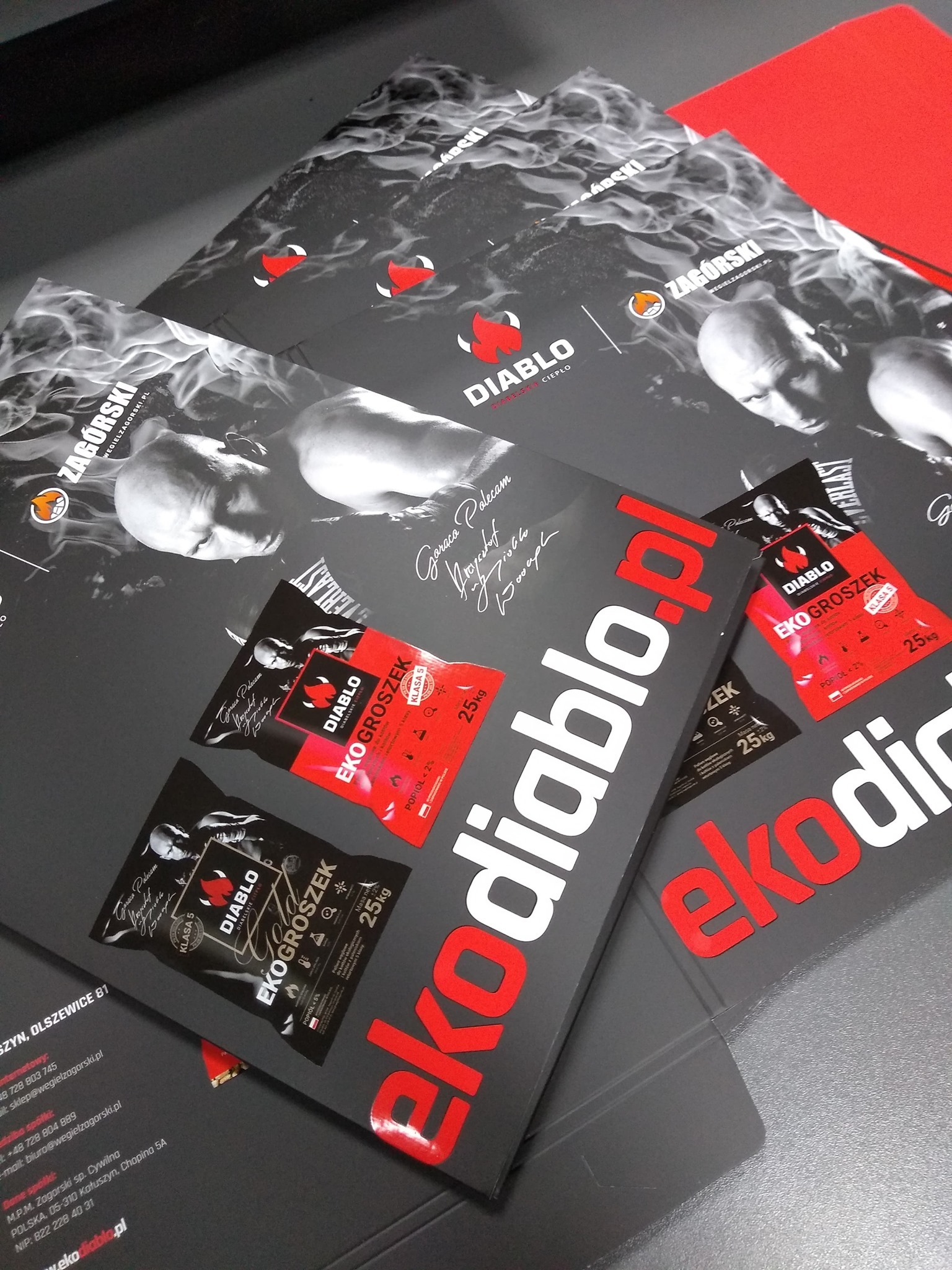 Stos czarnych, matowych ulotek i folderów reklamowych z czerwonymi akcentami, prezentujących markę 'ekodiablo.pl' i jej produkt 'Eko Groszek', widoczne logo i dane teleadresowe firmy.
