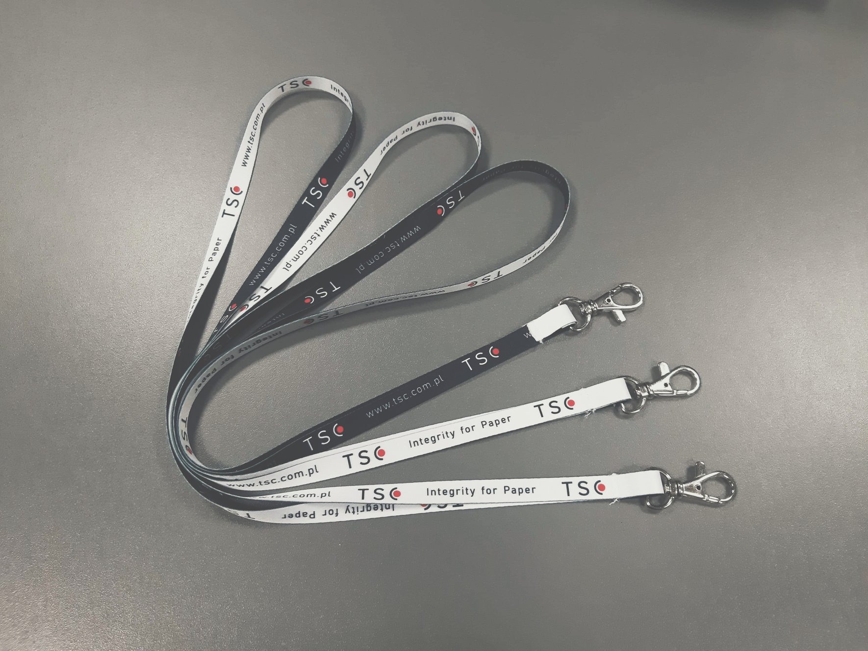 Trzy smycze reklamowe z karabińczykami, z logo firmy TSC, leżące na szarym tle.