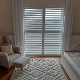 Białe shuttersy zamontowane w oknie, widoczne z wnętrza pokoju z beżowym dywanem w geometryczne wzory, kremowym fotelem i białymi zasłonami.