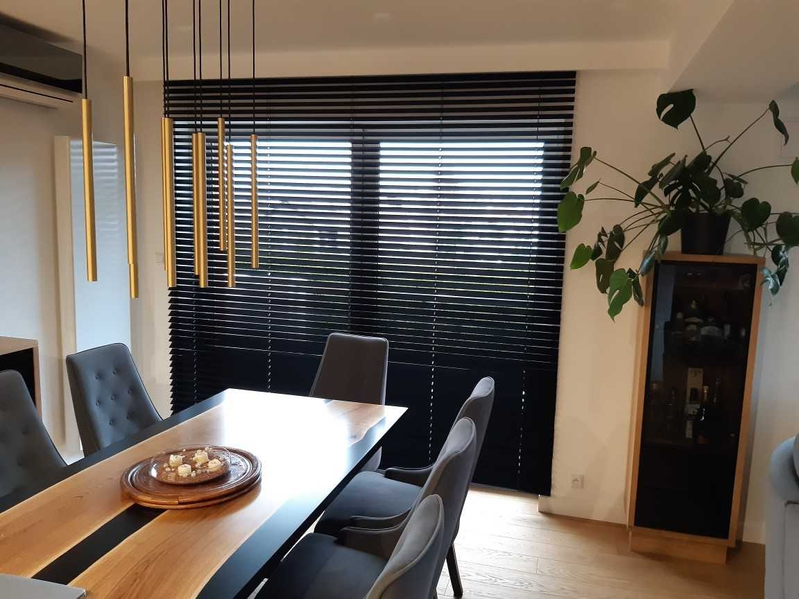 Czarne żaluzje drewniane z wąskimi lamelami zamontowane w oknie, widoczne z perspektywy jadalni z drewnianym stołem i nowoczesną lampą wiszącą.