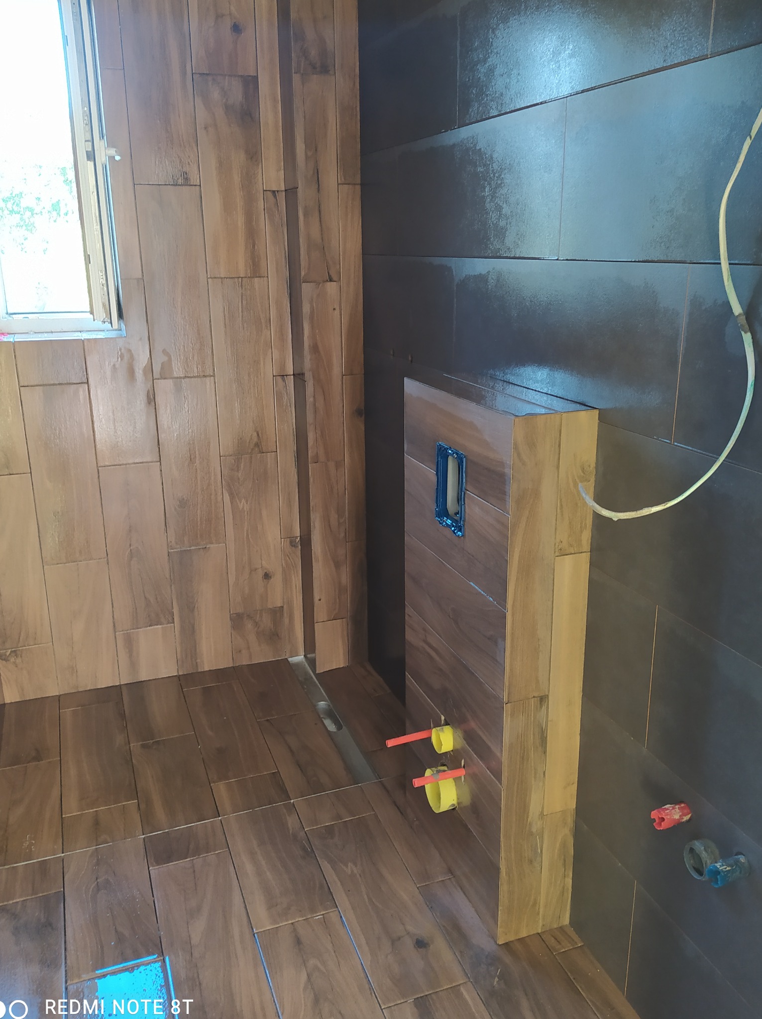 Wnętrze łazienki w trakcie prac wykończeniowych: drewnopodobne płytki na ścianach i podłodze, widoczne przyłącza wodne i elektryczne, surowy stan instalacji.