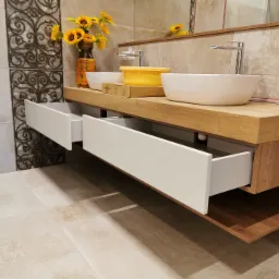 Łazienka z drewnianym blatem, dwiema białymi umywalkami nablatowymi, otwartymi szufladami i dekoracyjnym panelem ściennym z motywem ornamentów.